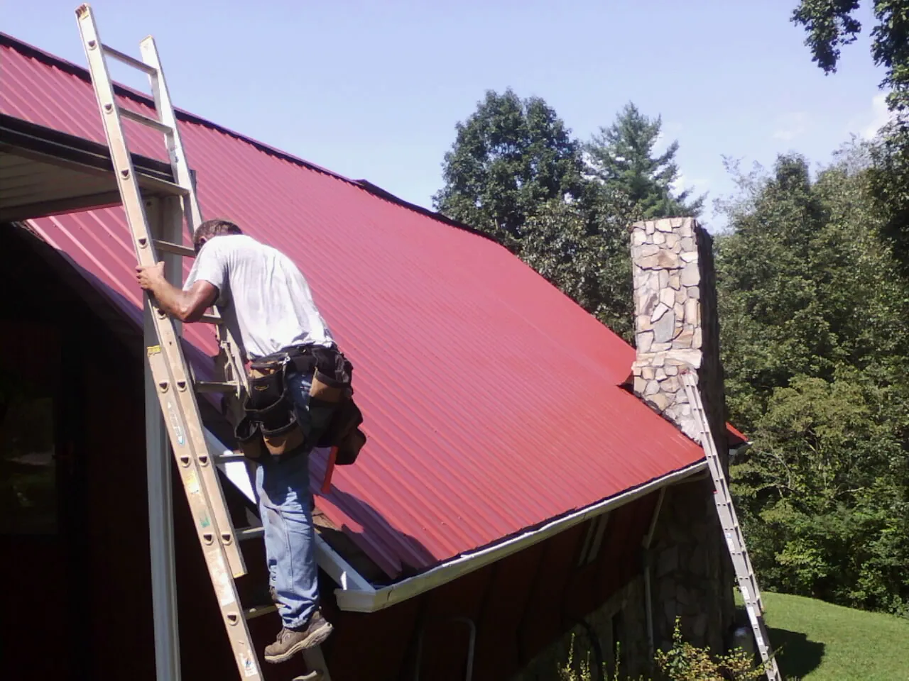 Roofer on a ladder beside a red metal roof and stone chimney — Soffit & Fascia Repair by Roofing Brothers in Little Falls